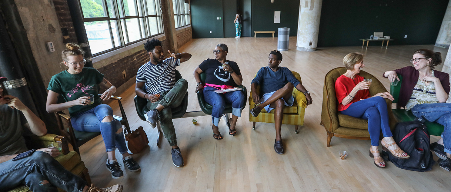 Seven people sitting in a line of chairs talking to each other.